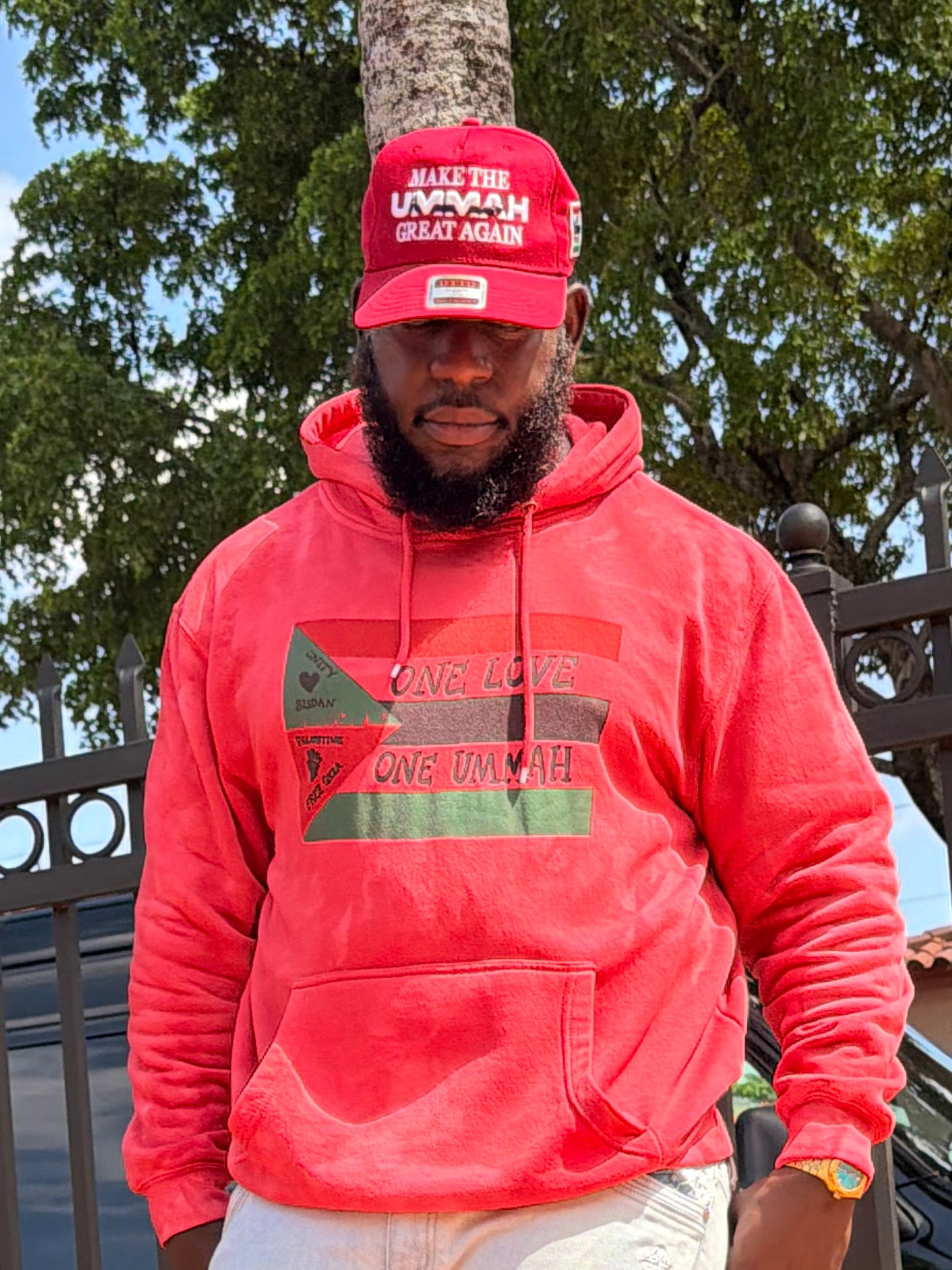 A man wearing a red hoodie with a Freedom Flagfthemed graphic and the words 'ONE LOVE ONE UMMAH' on the front, paired with a matching red cap that reads 'MAKE THE UMMAH GREAT AGAIN.' He is standing outdoors in front of trees and a fence.